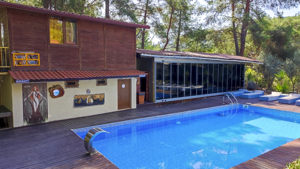 A serene poolside setting features a rectangular blue swimming pool bordered by wooden decking. Adjacent to the pool is a two-story wooden structure with large windows and a smaller section with artistic murals and a door. The area is surrounded by lush green trees and comfortable seating creates a relaxing environment.