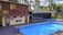 A serene poolside setting features a rectangular blue swimming pool bordered by wooden decking. Adjacent to the pool is a two-story wooden structure with large windows and a smaller section with artistic murals and a door. The area is surrounded by lush green trees and comfortable seating creates a relaxing environment.