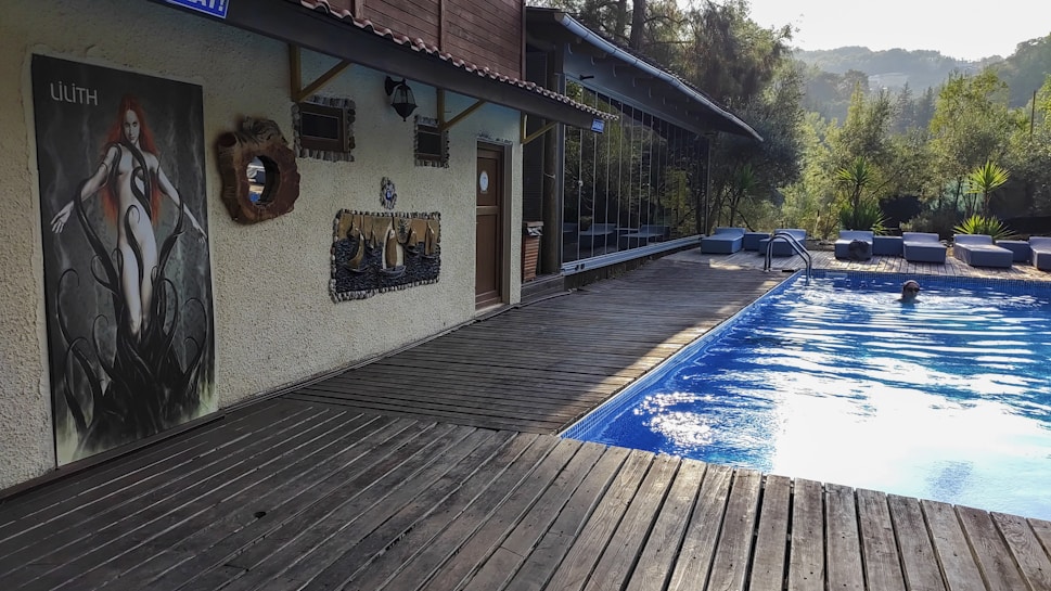A sunny outdoor pool area surrounded by lush greenery, featuring lounge chairs and a person swimming. The pool is bordered by a wooden deck, leading to a wall adorned with various decorations, including a large art poster.