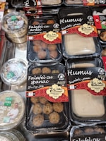 Various packaged Lebanese food items are displayed on a market shelf. Products include falafel with spinach, falafel with bell pepper, and different types of hummus, all from a brand called Zein. The packaging features red and black designs with both English and Arabic text. Another brand called Apetit Vegetarian is also visible, showcasing hummus varieties.