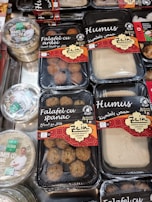 Various packaged Lebanese food items are displayed on a market shelf. Products include falafel with spinach, falafel with bell pepper, and different types of hummus, all from a brand called Zein. The packaging features red and black designs with both English and Arabic text. Another brand called Apetit Vegetarian is also visible, showcasing hummus varieties.