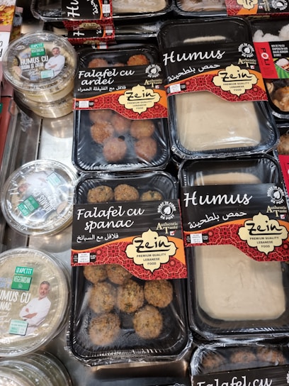 Various packaged Lebanese food items are displayed on a market shelf. Products include falafel with spinach, falafel with bell pepper, and different types of hummus, all from a brand called Zein. The packaging features red and black designs with both English and Arabic text. Another brand called Apetit Vegetarian is also visible, showcasing hummus varieties.
