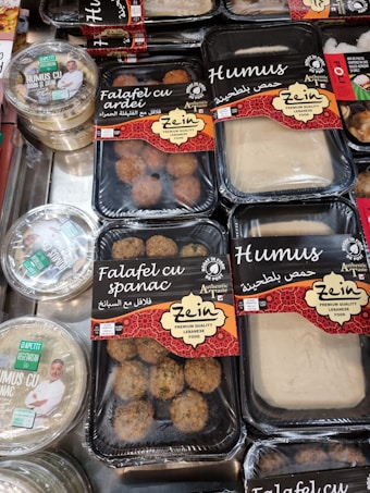 Various packaged Lebanese food items are displayed on a market shelf. Products include falafel with spinach, falafel with bell pepper, and different types of hummus, all from a brand called Zein. The packaging features red and black designs with both English and Arabic text. Another brand called Apetit Vegetarian is also visible, showcasing hummus varieties.