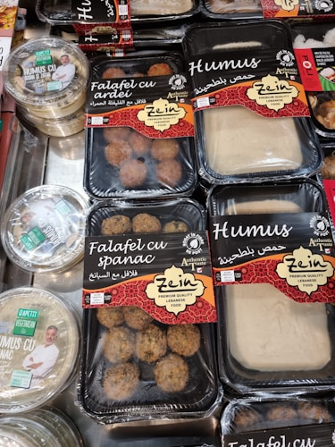 Various packaged Lebanese food items are displayed on a market shelf. Products include falafel with spinach, falafel with bell pepper, and different types of hummus, all from a brand called Zein. The packaging features red and black designs with both English and Arabic text. Another brand called Apetit Vegetarian is also visible, showcasing hummus varieties.
