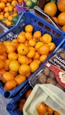 A display of fresh oranges in blue crates at a market or grocery store. Nearby, packaged falafel is visible with a label in multiple languages. The scene includes an abundance of bright, ripe oranges and a glimpse of other green produce in mesh packaging.