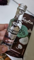 A small bottle of clear liquor labeled 'Stalinska Silver Premium Vodka' is held by a hand. Behind the bottle is a package of chocolate with coconut pieces, labeled as having 33% cacao and featuring coconut imagery.