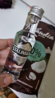 A small bottle of clear liquor labeled 'Stalinska Silver Premium Vodka' is held by a hand. Behind the bottle is a package of chocolate with coconut pieces, labeled as having 33% cacao and featuring coconut imagery.