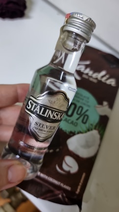 A small bottle of clear liquor labeled 'Stalinska Silver Premium Vodka' is held by a hand. Behind the bottle is a package of chocolate with coconut pieces, labeled as having 33% cacao and featuring coconut imagery.