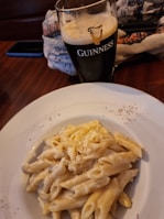 A glass of dark beer branded with the Guinness logo sits on a wooden table next to a white plate filled with creamy penne pasta garnished with grated cheese. A patterned cloth and a mobile phone in a dark case are partially visible in the background.