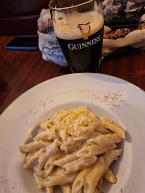 A glass of dark beer branded with the Guinness logo sits on a wooden table next to a white plate filled with creamy penne pasta garnished with grated cheese. A patterned cloth and a mobile phone in a dark case are partially visible in the background.