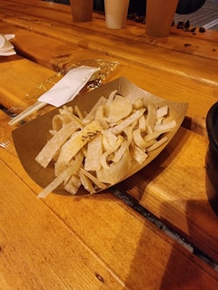 A wooden table with a cardboard container holding fried tortilla strips. A plastic fork and wrapped napkin are placed next to the container, and there are paper cups and miscellaneous items in the background.