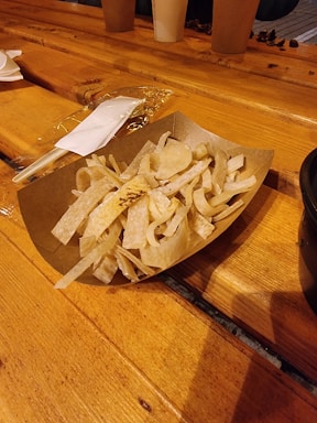 A wooden table with a cardboard container holding fried tortilla strips. A plastic fork and wrapped napkin are placed next to the container, and there are paper cups and miscellaneous items in the background.