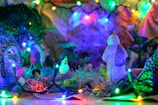 A vibrant and colorful scene with bright, multi-colored Christmas lights illuminating a miniature landscape. The focal point is a small, sculpted figure resembling a snowman, surrounded by greenery and other tiny decorative elements. The lights cast a warm, festive glow over the scene.