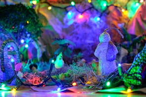 A vibrant and colorful scene with bright, multi-colored Christmas lights illuminating a miniature landscape. The focal point is a small, sculpted figure resembling a snowman, surrounded by greenery and other tiny decorative elements. The lights cast a warm, festive glow over the scene.