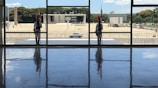 Two guards dressed in ceremonial uniforms stand at attention inside a building with large windows, with a view of a government building and a car parked outside. The floor reflects the figures, and trees and a clear sky can be seen in the background.