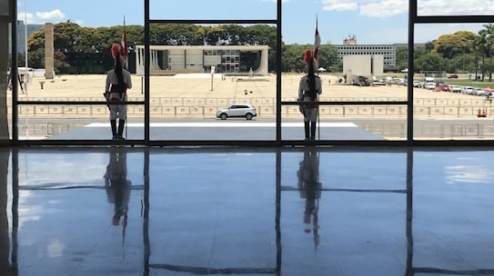 Two guards dressed in ceremonial uniforms stand at attention inside a building with large windows, with a view of a government building and a car parked outside. The floor reflects the figures, and trees and a clear sky can be seen in the background.