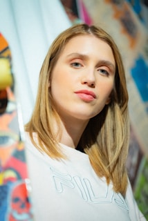 A person with light brown hair is wearing a white shirt with 'Fila' on it. The background is colorful and abstract, possibly graffiti or street art.