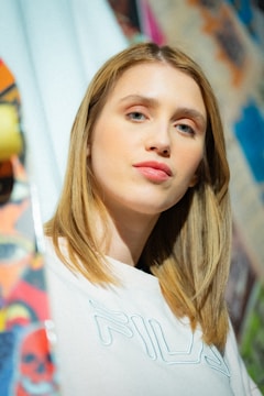 A person with light brown hair is wearing a white shirt with 'Fila' on it. The background is colorful and abstract, possibly graffiti or street art.