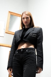 A woman with long blonde hair is wearing a dark cropped blazer adorned with a row of safety pins and black jeans. She stands in a minimalist setting with several gold-framed mirrors on the wall behind her, giving a modern and edgy look.