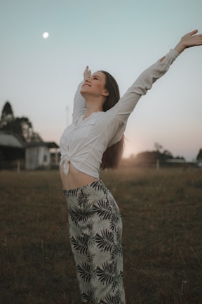 A woman standing in a field at sunrise, arms raised in joyful worship.