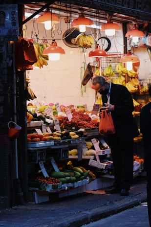 A man wearing a dark suit is shopping at a fruit stand filled with a variety of colorful fruits including bananas, grapes, and avocados. Warm lighting from multiple overhead lamps creates a cozy and inviting atmosphere. Prices are handwritten on small signs attached to the fruit displays.