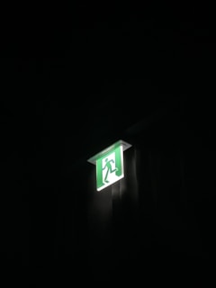 A glowing green emergency exit sign is mounted on a wall. The sign features a silhouette of a person running through a door, indicating the direction of the exit. It is illuminated against a dark background, highlighting its importance.