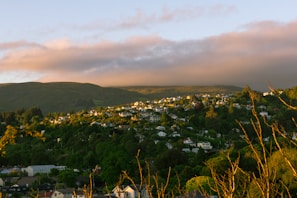 A scenic view of a hillside community with numerous houses nestled among lush greenery. The area is bathed in warm sunlight, creating a serene and picturesque atmosphere. The background features rolling hills under a sky covered with soft clouds.