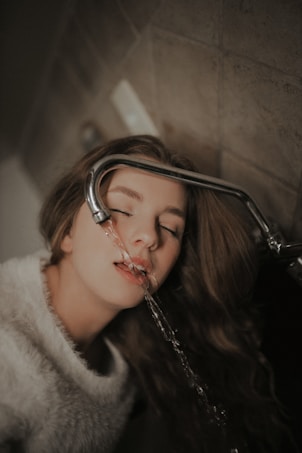 A person with long hair is drinking water from a tap with eyes closed, creating a serene atmosphere. The background is tiled, and the overall lighting is soft and warm.
