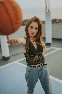 A young woman stands on an outdoor basketball court, extending her arm with a basketball balanced on her finger. She wears a black lace top and blue jeans, looking directly into the camera with a confident expression. The background shows a fence and support poles, suggesting an urban rooftop setting.