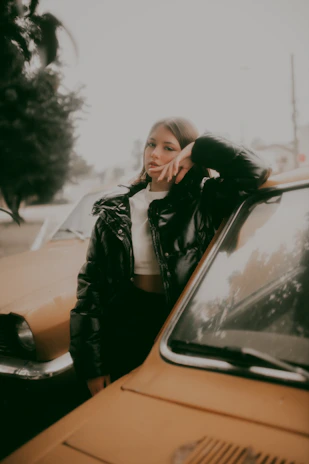 A man in a sharp black blazer leaning against a vintage car under soft evening light.