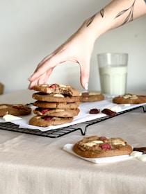 A hand with a tattoo reaches for a stack of homemade cookies on a cooling rack. The cookies are topped with white and dark chocolate chunks along with dried red fruit. A glass of milk sits in the background on a beige tablecloth, contributing to a cozy, homey atmosphere.