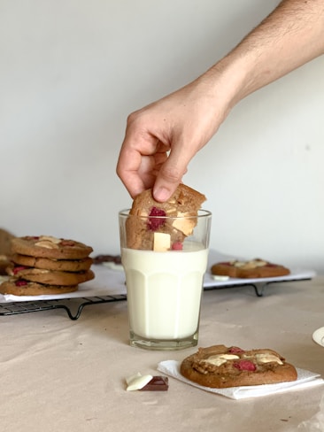 A hand is dipping a cookie loaded with white and red chunks into a tall glass of milk. Several cookies sit on a cooling rack in the background, accompanied by visible chunks of chocolate on the table.