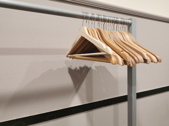 A series of wooden coat hangers are neatly aligned on a metal clothing rack, against a plain wall with a shadow cast by the hangers. The hangers are angled in different directions, creating a visually interesting pattern.