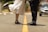 A couple stands face to face on an empty road. The woman is wearing a white dress and black high-top sneakers and has one foot playfully lifted behind her. The man is dressed in dark jeans and black sneakers. The road is lined with trees and a blurred car is visible in the background.