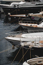 Several luxury yachts are docked in a marina, with their sleek designs and polished surfaces. The yachts are lined up closely, each tied with ropes to the dock. The water reflects the hulls of the boats, creating a calm and serene atmosphere.