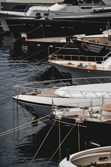 Several luxury yachts are docked in a marina, with their sleek designs and polished surfaces. The yachts are lined up closely, each tied with ropes to the dock. The water reflects the hulls of the boats, creating a calm and serene atmosphere.