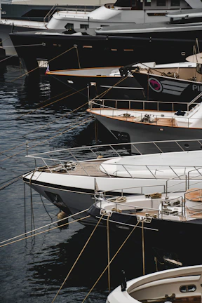 Several luxury yachts are docked in a marina, with their sleek designs and polished surfaces. The yachts are lined up closely, each tied with ropes to the dock. The water reflects the hulls of the boats, creating a calm and serene atmosphere.