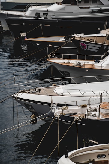Several luxury yachts are docked in a marina, with their sleek designs and polished surfaces. The yachts are lined up closely, each tied with ropes to the dock. The water reflects the hulls of the boats, creating a calm and serene atmosphere.