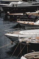 Several luxury yachts are docked in a marina, with their sleek designs and polished surfaces. The yachts are lined up closely, each tied with ropes to the dock. The water reflects the hulls of the boats, creating a calm and serene atmosphere.