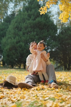 A parent and child sit together on a blanket in a park filled with fallen yellow leaves. The child, dressed in a pink outfit, is being held and looks cheerful as they point towards the sky. Surrounding them are tall, green trees and more autumn leaves.