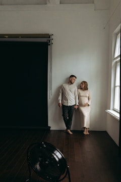 A couple stands closely together, leaning against a white wall in an interior setting. The woman is dressed in a form-fitting, light-colored dress and gently embraces her belly, suggesting pregnancy. The man wears a casual light shirt and pants, looking at the woman warmly. The wooden floor, natural light from a window, and a black photography backdrop create an intimate atmosphere.
