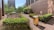 A rooftop garden with neatly arranged flower beds bordered by green hedges. In the foreground, there are two wooden bee hives or boxes. The area is surrounded by large buildings, and sunlight casts shadows on the walls. Trees and bushes are visible in the background.