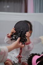 A child dressed in white and pink pajamas is holding up a decorative black object to their face. A hand is reaching towards the child, possibly trying to touch or take the object. The background is soft and out of focus, suggesting an indoor setting, possibly a bedroom or living room.