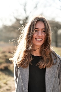 A smiling woman with long hair standing outdoors in a natural setting, with sunlight illuminating her hair and creating a warm atmosphere. The background is slightly blurred, enhancing the focus on her expression and presence.