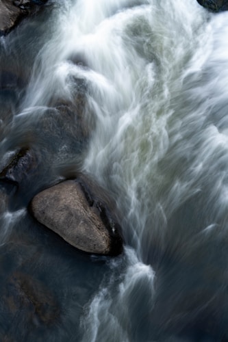 Water flows over rocks in a stream with a smooth, blurred motion. The water appears dynamic and energetic, cascading around the stones.