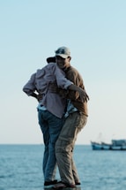 Two people are embracing on a small platform by the sea, with boats visible in the background. The sky is clear and the scene conveys a moment of friendship or companionship.