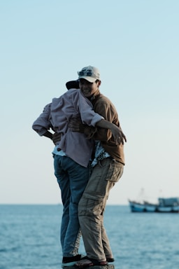 Two people are embracing on a small platform by the sea, with boats visible in the background. The sky is clear and the scene conveys a moment of friendship or companionship.