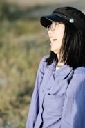A person with dark hair, wearing glasses and a black cap, is laughing joyfully. They are dressed in a light purple shirt and are outdoors with a blurred background of greenery.