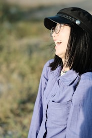 A person with dark hair, wearing glasses and a black cap, is laughing joyfully. They are dressed in a light purple shirt and are outdoors with a blurred background of greenery.
