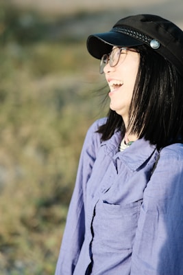 A person with dark hair, wearing glasses and a black cap, is laughing joyfully. They are dressed in a light purple shirt and are outdoors with a blurred background of greenery.
